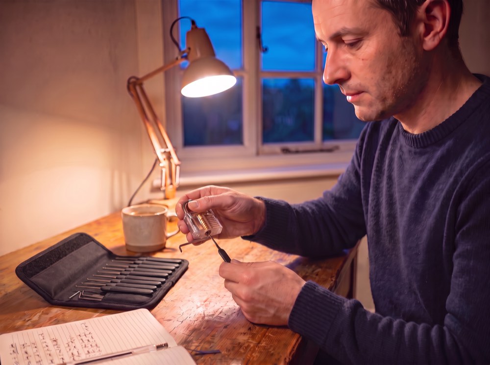A man at a lamp-lit desk picking a clear practice padlock with a hook pick and tension wrench, his Sparrows pouch open beside him